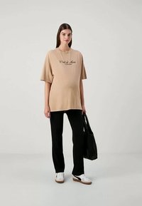 Gravid kvinne som har på seg en beige oversized T-skjorte med tekst, svarte bukser, hvite joggesko, og holder en svart håndveske mot en ensfarget bakgrunn.