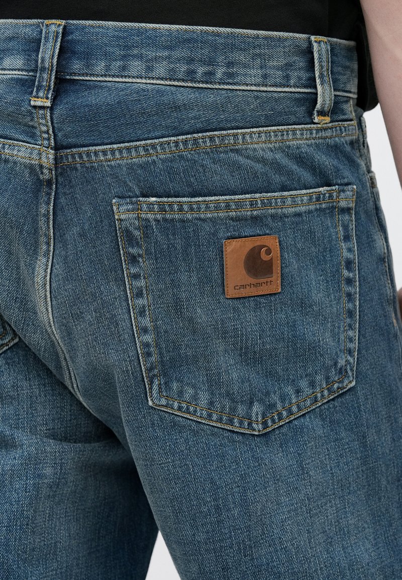 Blue denim jeans featuring a back pocket with a brown leather Carhartt logo patch, stitching details, and a classic straight-leg design.