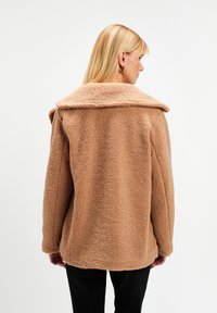 Kobieta z włosami blond do ramion, ubrana w jasnobrązową kurtkę polarową i czarne spodnie, stojąca tyłem na tle jednolitej białej ściany.