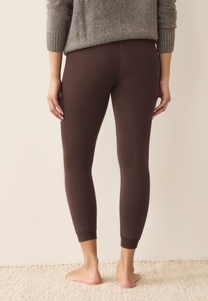 Naine seisab paljajalu tekstuurse vaiba peal, kandes pruune hästi istuvaid retuuse ja halli kootud kampsunit, näidatud tagantpoolt käed külgedel lõdvalt.