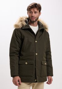 Uomo che indossa un parka invernale verde oliva con cappuccio foderato in pelliccia, frontale con bottoni e cerniera, e tasche a patta sopra un maglione bianco.