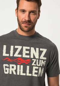 Šedé tričko s krátkým rukávem, s roztrhaným bílým nápisem "LIZENZ ZUM GRILLEN" a červeným plamenem na přední straně. Měkký materiál z bavlněné směsi.