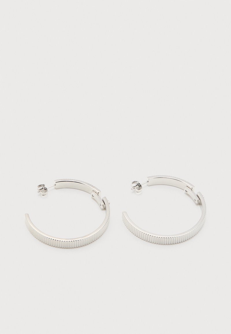 Σκουλαρίκια ημικυκλικού σχήματος με ασημί τόνο, με ραβδωτή υφή και πίσω μπαγιονέτες, σε λευκό φόντο.