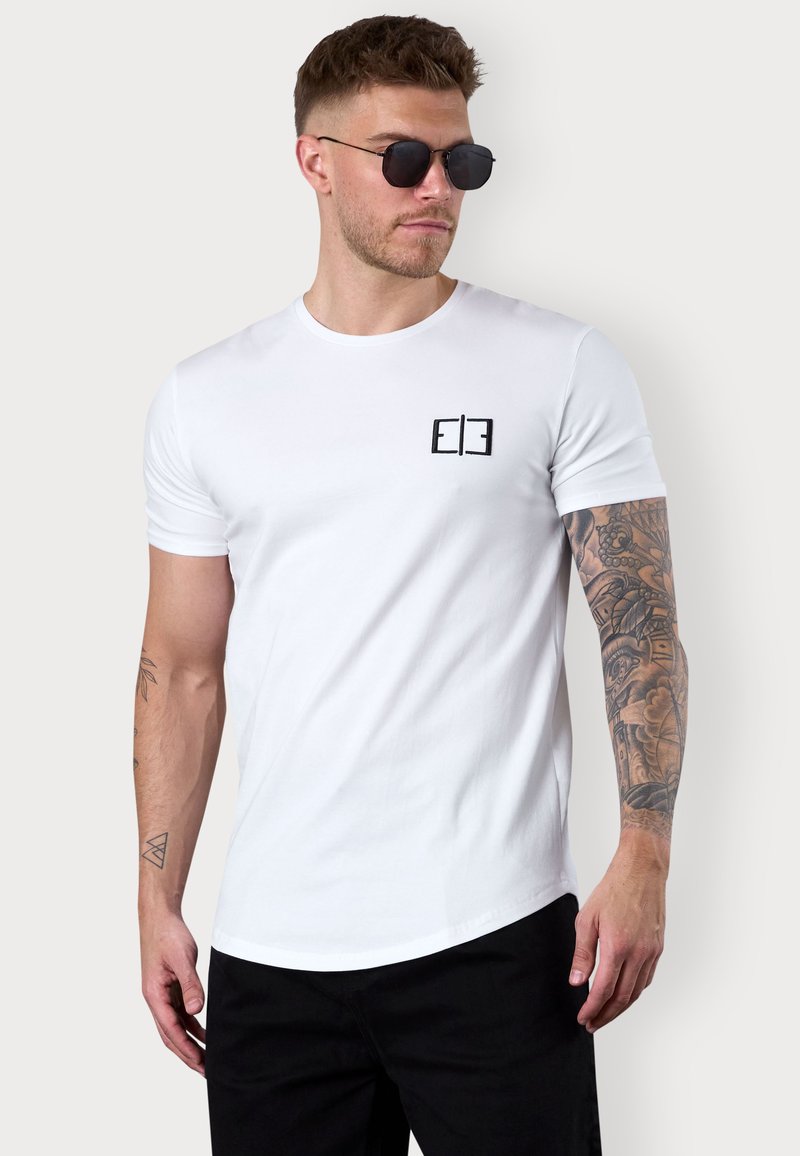 Weißes T-Shirt mit kurzen Ärmeln aus Baumwolle, versehen mit einem kleinen schwarzen Logo auf der Brust, kombiniert mit schwarzen Shorts. Strukturiertes Tattoo sichtbar am Arm.