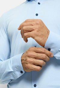 Chemise bleu clair à manches longues avec une texture douce. Comprend des boutons bleu foncé et un poignet avec une finition détaillée. Des mains ajustent le poignet.