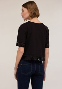 Camiseta negra corta con mangas cortas, que presenta detalles de lentejuelas negras brillantes en las mangas y el dobladillo, combinada con jeans azul oscuro.