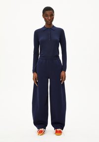 Frau in einem marineblauen Langarmpolo, weitgeschnittenen marineblauen Hosen und orangefarbenen Turnschuhen, stehend vor einem weißen Hintergrund.
