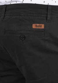 Schwarze Baumwollhose mit einer hinteren Tasche mit einem Knopf und einem braunen Lederetikett, auf dem "!Solid" steht. Glatte Textur und einfaches Design.
