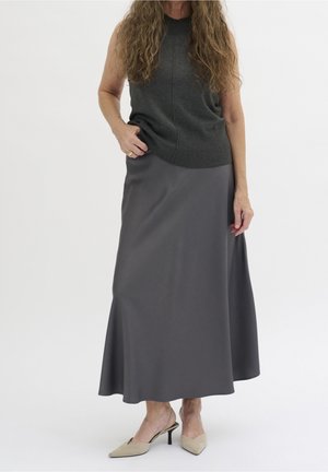 Kobieta ubrana w ciemnoszary bezrękawnik, długą szarą spódnicę i beżowe sandały na niskim obcasie typu slingback, stojąca na jednolitym tle.
