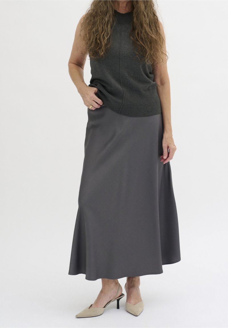 Femme portant un haut gris foncé sans manches, une longue jupe grise et des chaussures slingback beiges à petit talon, debout devant un fond uni.