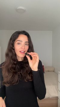 Femme aux cheveux longs tenant un petit appareil noir près de la bouche, debout dans une chambre avec un lit et une commode en arrière-plan.