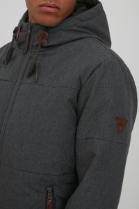 Men's gray padded jacket with a textured finish, hood, brown leather accents, and zippered pockets. Features adjustable drawstrings.