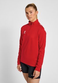Haut de sport rouge à manches longues avec col montant, zip à quart, et détail de logo blanc, associé à un short noir et à une texture de tissu lisse.