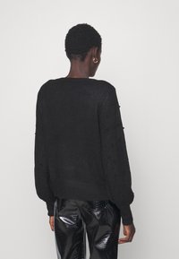 Maglione nero con maniche a sbuffo in tessuto testurizzato, dettagli in perline, scollo e orlo a coste; abbinato a pantaloni neri lucidi.