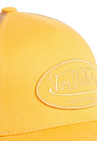 Casquette jaune en tissu texturé, arborant un patch logo brodé. Le design présente une visière courbée et structurée avec des coutures détaillées.