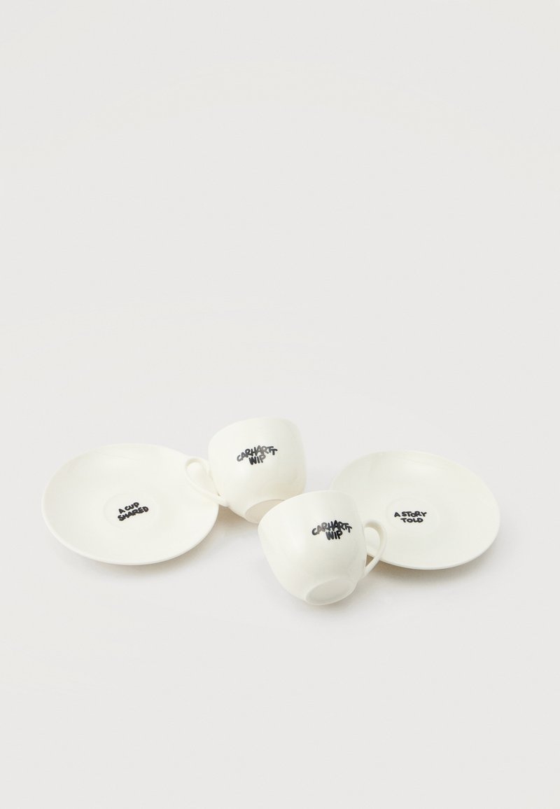 Zwei weiße Keramiktassen mit dem Text "CARHARTT WIP" und zwei weiße Untertassen mit "A CUP SHARED" und "A STORY TOLD" in schwarzer Schrift auf einfarbigem Hintergrund.