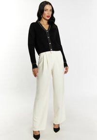 Schwarze Strickjacke mit Perlenakzenten und V-Ausschnitt, kombiniert mit hochgeschnittenen, weiten cremefarbenen Hosen und schwarzen spitzen Absätzen.