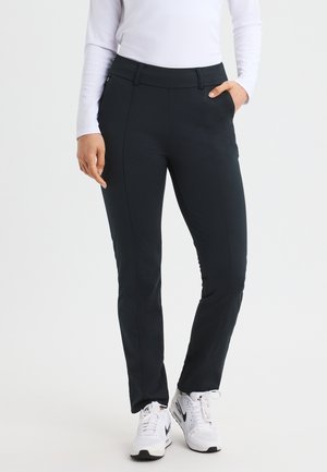 Zwarte, aangesloten broek met een gladde textuur, voorzien van voorzakken en subtiele stikdetails, gedragen met witte sportschoenen.