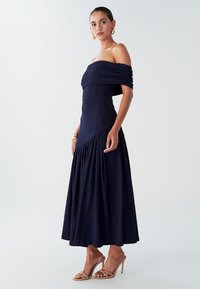 Robe bleu marine à épaules dénudées en tissu texturé, corsage ajusté et jupe fluide. Comprend des détails plissés et une couture diagonale.