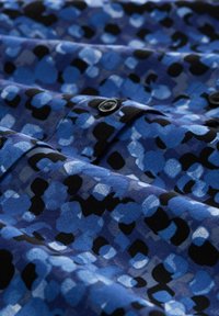 Blau gemusterter Stoff mit abstrakten Formen in verschiedenen Blautönen und Schwarz, mit einer glatten Textur und sichtbaren schwarzen Knöpfen.