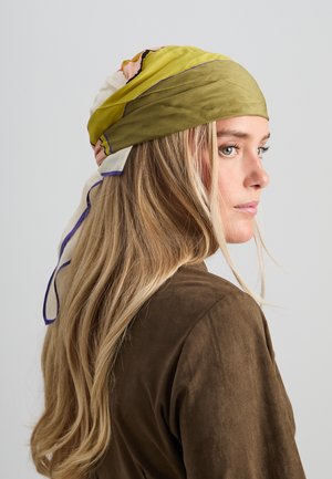 Jauna moteris su ilgais blondiniais plaukais, dėvinti žalią ir baltą galvos skarelę bei rudą striukę, atsukta į dešinę prieš pilką, vienspalvį foną.