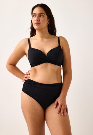 Pitkätukkainen nainen mustassa kaksiosaisessa uimapuvussa, katse sivulle, toinen käsi lantiolla, yksinkertaisen taustan edessä.