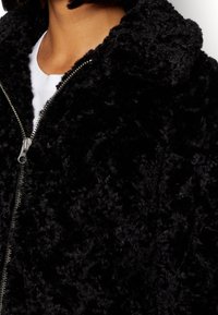 Svart syntetpälsjacka med dragkedja, med en hög krage och en mjuk, texturerad yta. Silverfärgade detaljer.