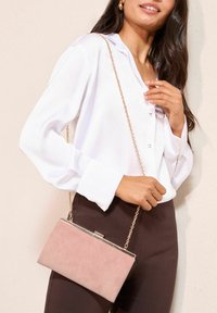 Prašnjavo roza plisirani kožnati neseser sa zlatnim lancem i metalnim kopčom, pravokutnog oblika, drži ga osoba u bijeloj bluzi i smeđim hlačama.