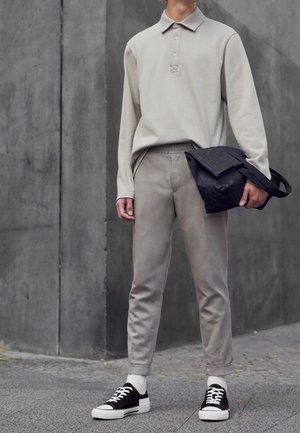 Person trägt beige langärmeliges Polo und Hose, hält eine schwarze Tasche, mit schwarz-weißen Sneakers auf einem grauen städtischen Gehweg stehend.