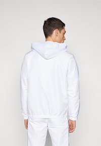 Chaqueta blanca con capucha, hecha de tejido suave, que presenta una cinturilla y puños elásticos, con acentos azules a lo largo de las mangas. Vista trasera sencilla.