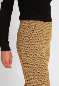 Un haut long à manches noires côtelé à côté de pantalons moulants marron et jaunes à motif géométrique et texture lisse.