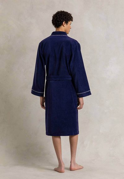 Νεαρό άτομο που στέκεται ξυπόλητο, φορώντας μπλε σκούρο μπουρνούζι μέχρι το γόνατο με λευκές λεπτομέρειες και ζώνη στη μέση, με την πλάτη γυρισμένη.