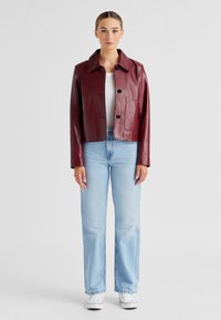 Burgunderrote Lederjacke mit Kragen, vorderen Knöpfen und zwei aufgesetzten Taschen. Kombiniert mit hellblauen, geraden Jeans und weißen Turnschuhen.