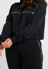 Svart kort hoodie med hög krage och långa ärmar, prydd med en vit "Nike Air"-logga på bröstet. Kombinerad med svarta leggings.