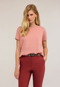 Růžové tričko s krátkým rukávem a kulatým výstřihem, spárované s červenými těsnými kalhotami. Oblečení doplňuje tenký, tmavý pásek se stříbrnou sponou.