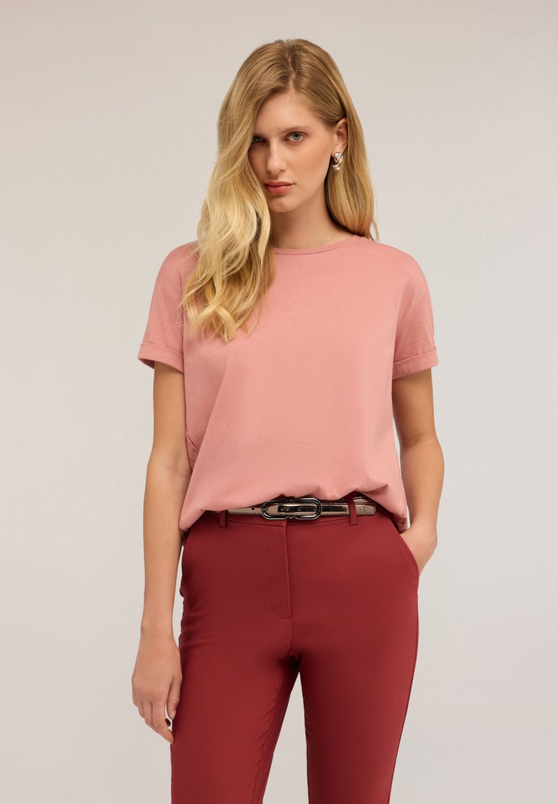 Růžové tričko s krátkým rukávem a kulatým výstřihem, spárované s červenými těsnými kalhotami. Oblečení doplňuje tenký, tmavý pásek se stříbrnou sponou.