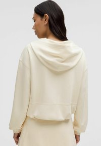 Felpa color crema con una texture morbida, vestibilità rilassata e cappuccio con coulisse. Presenta maniche ampie e orlo corto.