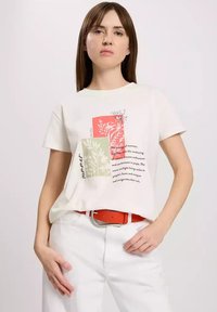 Kremowa koszulka z nadrukiem botanicznym w czerwono-zielonych kolorach; ma krótkie rękawy i luźny krój. W połączeniu z białymi spodniami i pomarańczowym paskiem.