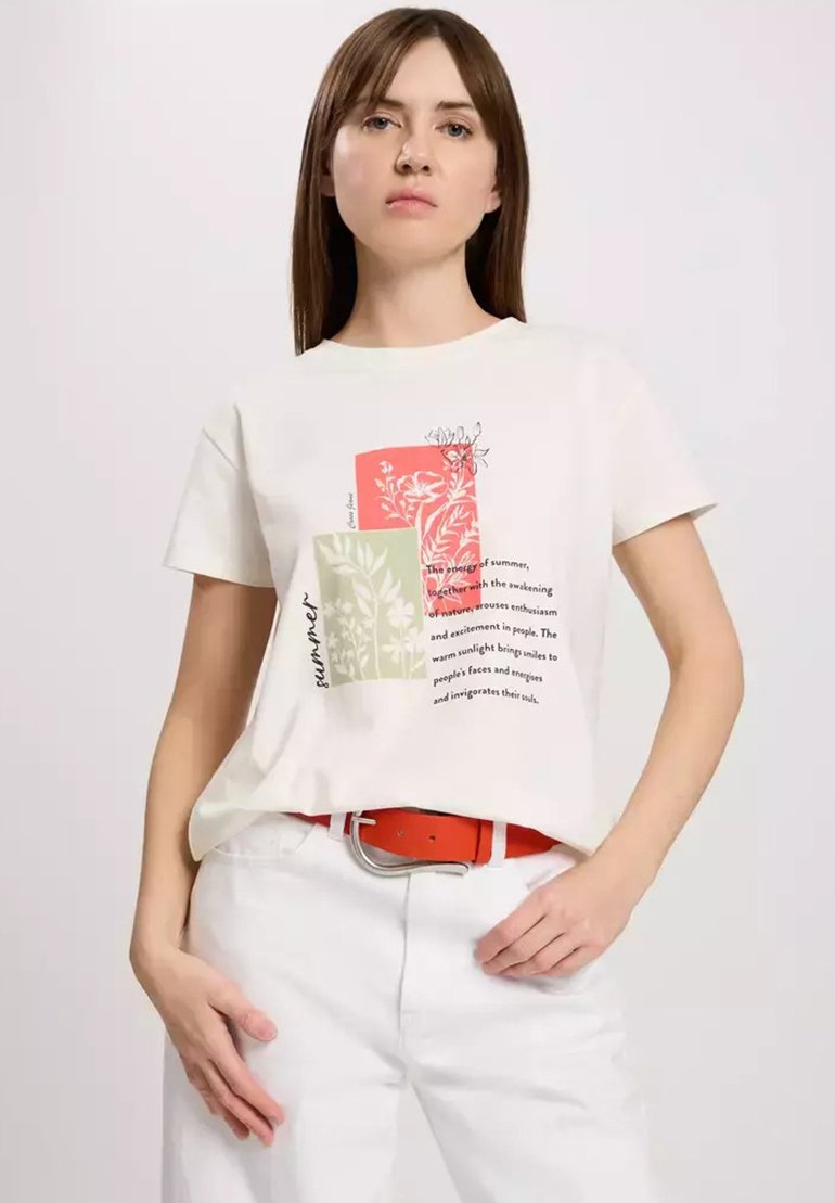 Kremowa koszulka z nadrukiem botanicznym w czerwono-zielonych kolorach; ma krótkie rękawy i luźny krój. W połączeniu z białymi spodniami i pomarańczowym paskiem.