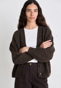 Bruine gebreide cardigan met een geribde halslijn, twee knopen en losse mouwen, gedragen over een witte top en gecombineerd met donkere broeken.