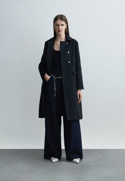 Γυναίκα που φοράει μαύρο παλτό με χρυσά κουμπιά, μαύρο τοπ, μπλε σκούρες παντελόνες καμπάνα με ζώνη αλυσίδα χρυσού, και λευκά μυτερά παπούτσια, όρθια.