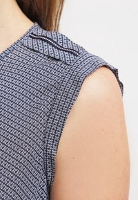 Mörkblå ärmlös blus med ett geometriskt vitt mönster, klassisk krage och textil med en slät yta.