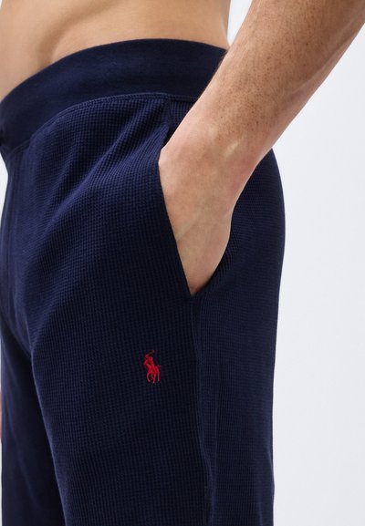 Χέρι που ξεκουράζεται μέσα στην τσέπη σκούρου μπλε παντελονιού με υφή, με κόκκινο κεντημένο λογότυπο αλόγου στον μηρό.