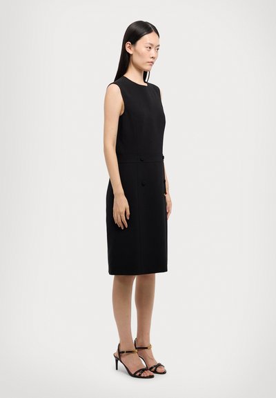 Schwarzes ärmelloses Kleid mit rundem Ausschnitt, zwei vorderen Knöpfen und geradem Schnitt. Getragen mit schwarzen, hochhackigen Sandalen.
