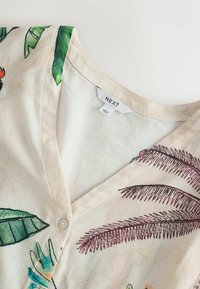 Krém színű V-nyakú blúz zöld és barna trópusi leveles mintával, elöl egyetlen fehér gombbal, a gallér belsején „NEXT” felirattal.