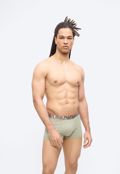 Μυώδης άνδρας με κοτσίδες στέκεται γυμνόστηθος φορώντας ανοιχτοπράσινα εσώρουχα Emporio Armani μπροστά σε απλό λευκό φόντο.