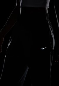 Zwarte sportbroek met een elastische tailleband. Voorzien van een reflecterend witte Nike-logo op de dij, tegen een donkere achtergrond.