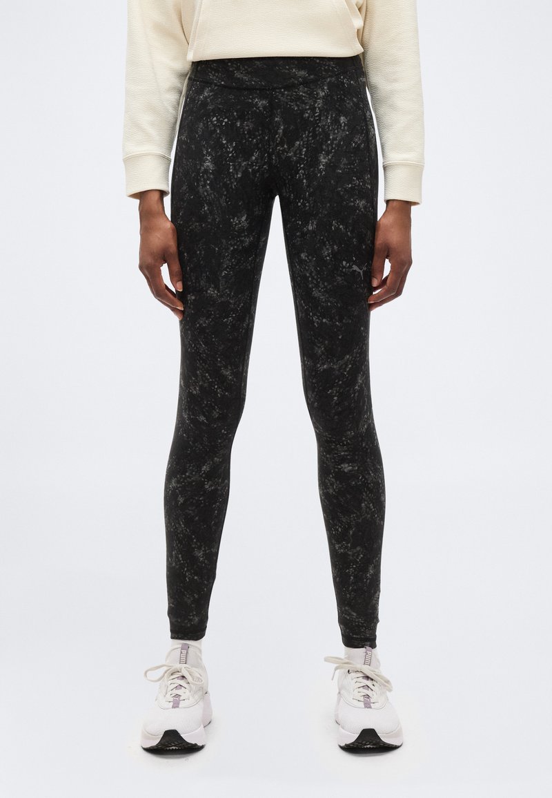 Άτομο που φορεί μαύρο μάρμαρο κολάν, εκρού φούτερ και λευκά αθλητικά παπούτσια Puma, στέκεται μπροστά σε ένα απλό λευκό φόντο.