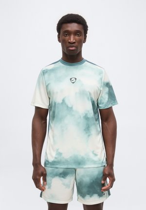 Homme debout face à l’avant portant un t-shirt de sport vert et blanc à motif de nuages et un short assorti sur un fond uni.