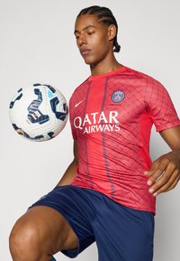 Rød kortærmet fodboldtrøje med diamantmønster og "QATAR AIRWAYS" logo. Blå shorts, spiller holder en multirød fodbold.